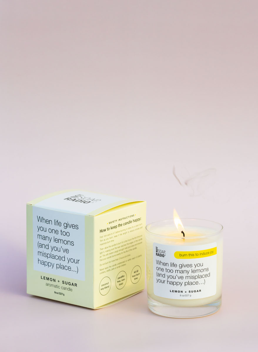 A lemon scented candle that has been lit next to the product's box with the label showing on top of a light pink background.