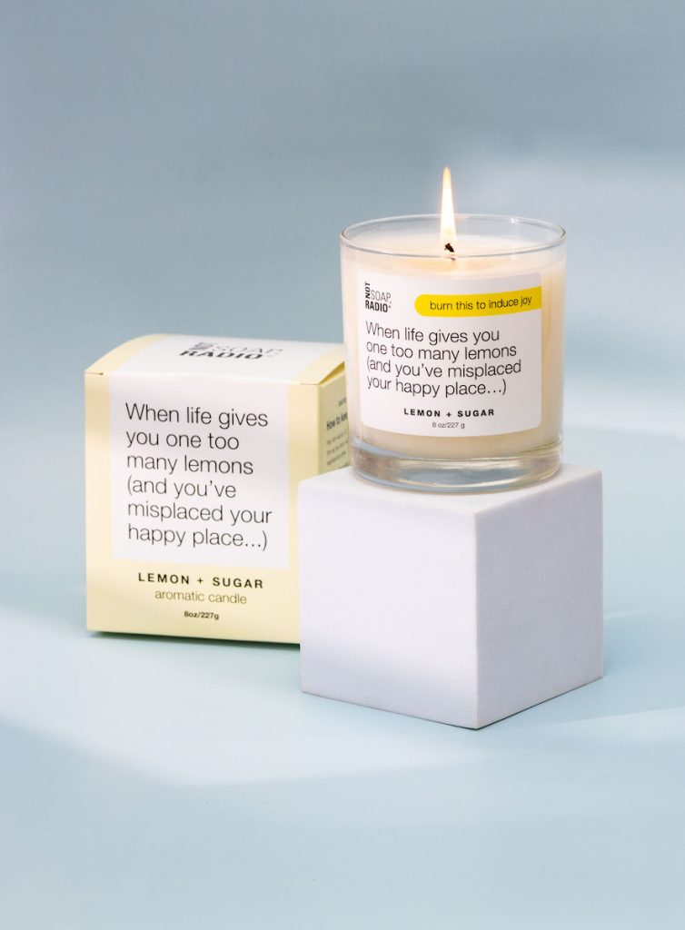 Staged lit candle on top of foam white cube with the product's packaging to the left on a light blue background. Product label and packaging designed by Seaweed Studio, as well as the photography