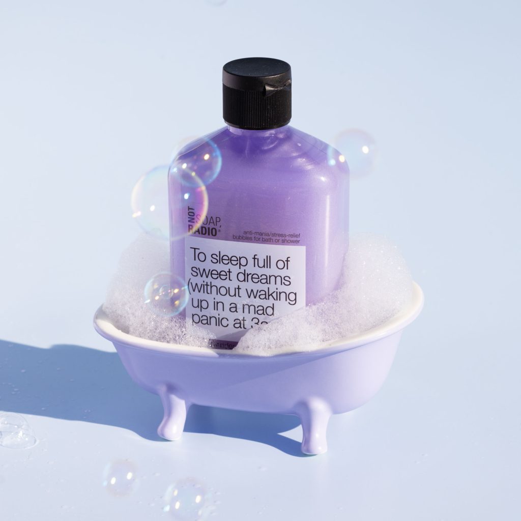 Purple body wash bottle sitting inside of a small soapy bathtub with light bubbles floating around the photo.