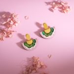 A pair of pink and green feathered earrings diagonally arranged on top of a light pink background with pink flower petals scattered around.